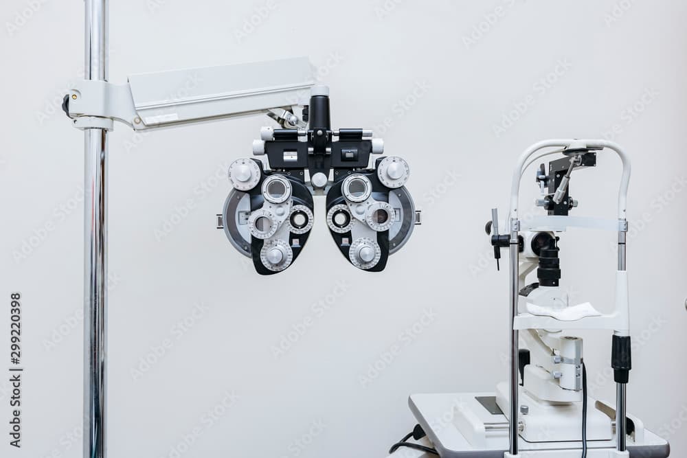 A clean wide shot of an eye exam room: a phoropter on its swing arm against a white wall beside a slit lamp on a sliding table. No people in frame, an uncluttered establishing shot of the room itself.