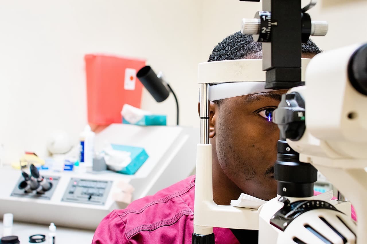 A patient resting their chin and forehead on a slit lamp biomicroscope while a clinician prepares to measure eye pressure. Optometry consumables and a sharps bin sit softly out of focus in the background of the exam room.