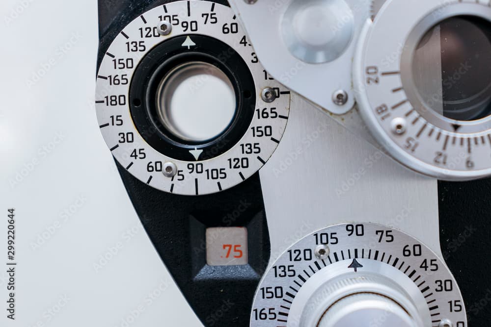 An extreme close-up of a phoropter's refraction dials, showing the engraved axis scales, numbered cylinder wheels, and a small window reading a power value. Fine mechanical detail that generative models routinely get wrong.