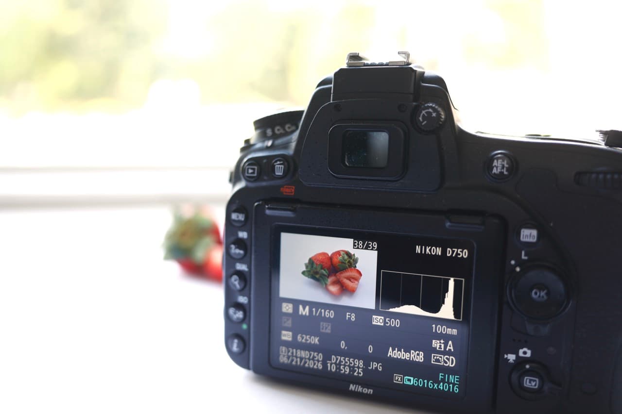 A Nikon D750 resting on a bright windowsill, its rear screen showing the strawberry shot alongside a healthy histogram and the capture settings (Manual, 1/160, f8, ISO 500, 6250K). The actual strawberries sit softly out of focus behind the camera, lit by the window.