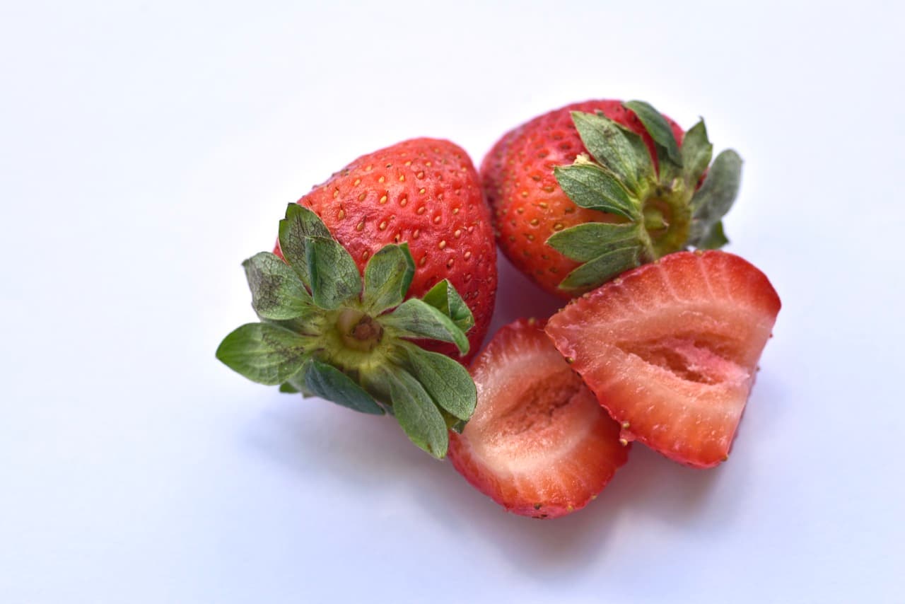 The finished strawberry still life as a straight-out-of-camera JPEG with white balance set correctly: clean neutral white background, true-to-life red fruit and green leaves. Two whole strawberries and one halved, lit softly from the window.