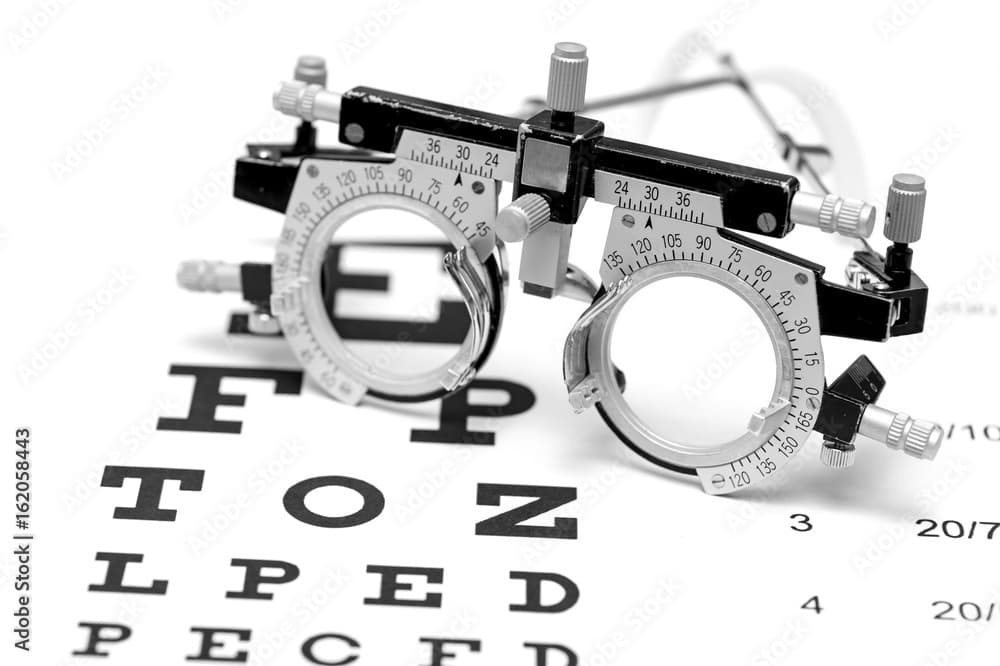 A black-and-white close-up of an optometry trial frame resting on a printed Snellen eye chart, the adjustable lens cells and rotation scales in sharp focus over rows of test letters.