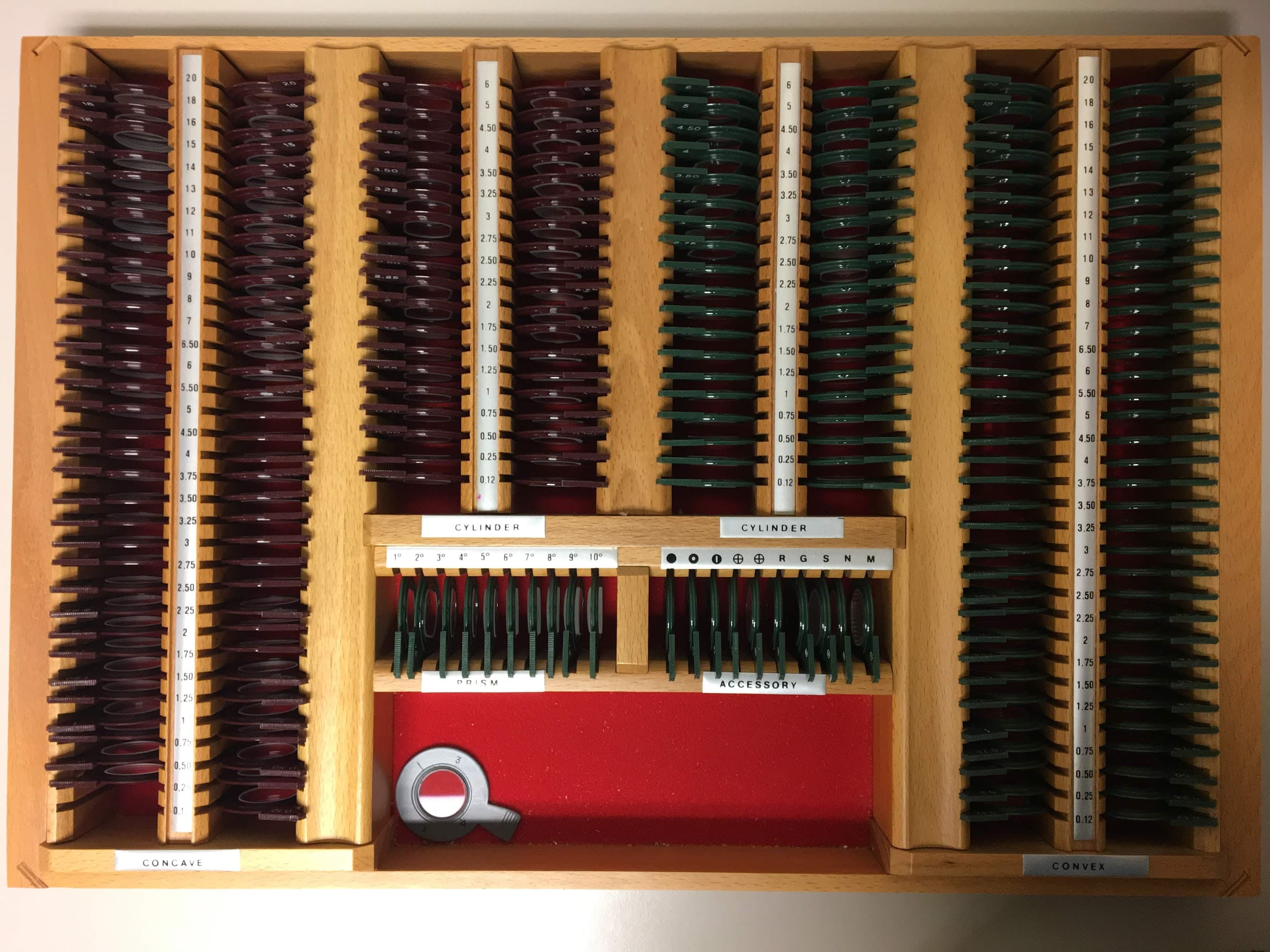 A top-down view of an open wooden optometry trial lens set, rows of concave and convex lenses and cylinder lenses seated in labeled trays, with a lens holder in the center well. An uncommon subject a vision model may not recognize on sight.